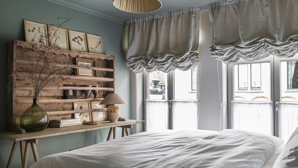 Ein Schlafzimmer mit einem Bett, einer Holzregal mit verschiedenen Gegenständen, einer Lampe und einem Fenster mit Vorhängen.