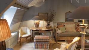 Ein gemütliches Dachzimmer mit geneigter Decke, einem Holztisch mit Lampe, einer Couch mit Kissen und einem Sessel.