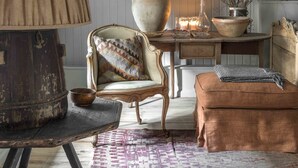 Ein gemütliches Zimmer mit Holzbalkendecke, einer großen Lampe, einem Vintage-Stuhl, einem kleinen Tisch mit Kerzen und einem gemusterten Teppich.