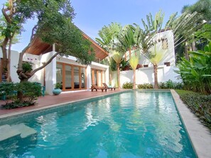 Zona de piscina con una piscina azul cristalina, rodeada de palmeras y un edificio blanco con amplias puertas de cristal.