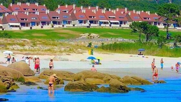 A coastal resort with multiple red-roofed buildings, a sandy beach, and rocky areas with people enjoying the water. - Complejo Turístico Raeiros (O Grove)