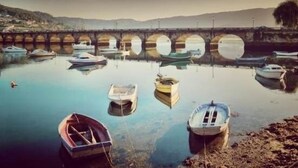 A calm harbor with several boats docked, a bridge in the background, and a hilly landscape. - Complejo Turístico Raeiros (O Grove)