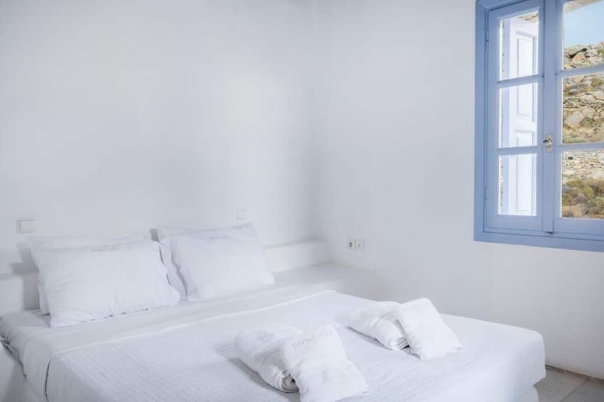 Una habitación blanca con una cama, una ventana que da a rocas y una puerta.