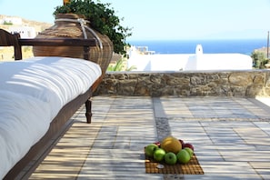 Un lit avec vue sur la mer, un mur en pierre et une table garnie de fruits.