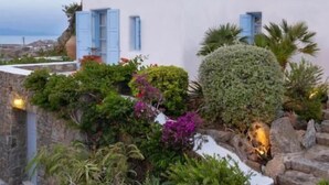 A white building with blue shutters, surrounded by lush greenery and a stone pathway. - Nama Villas (Mykonos)