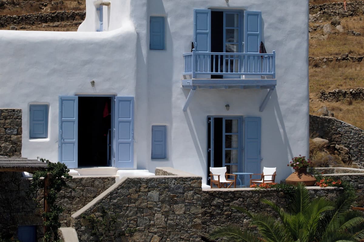 Un edificio blanco con contraventanas azules y un balcón, rodeado de muros de piedra y vegetación.