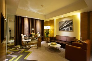 A hotel room with a sofa, armchairs, a coffee table, a desk with a chair, a lamp, a flower arrangement, and a framed picture on the wall.