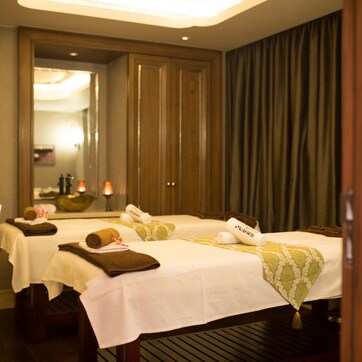 A hotel room with two beds, each with a white bedspread and a green patterned throw. There are towels and a small table with a bowl and a candle on it.