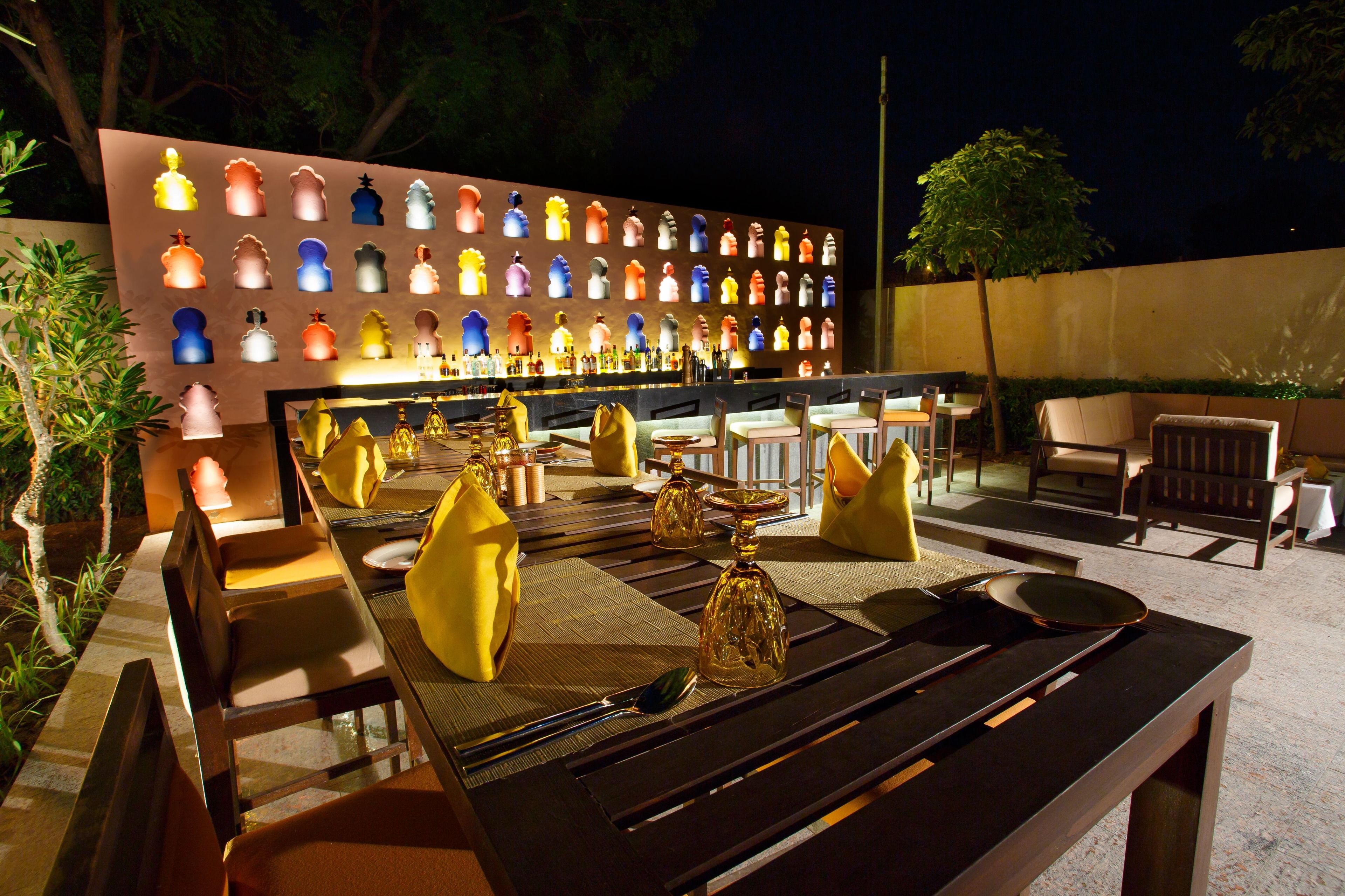 Un área de comedor al aire libre con una larga mesa dispuesta para dos personas, rodeada de sillas. Se ve un muro con piezas de ajedrez iluminadas al fondo.