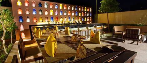 Un área de comedor al aire libre con una larga mesa dispuesta para dos personas, rodeada de sillas. Se ve un muro con piezas de ajedrez iluminadas al fondo.