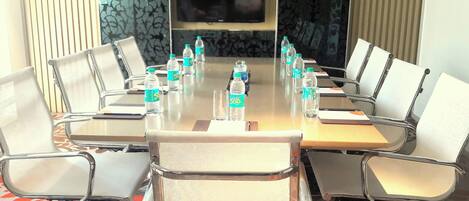 Un salón de conferencias con una larga mesa, sillas, botellas de agua y una pantalla de televisión.