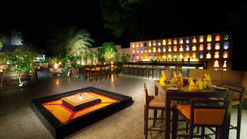 Un área de comedor al aire libre de noche con una chimenea, mesas, sillas y una barra con botellas iluminadas.