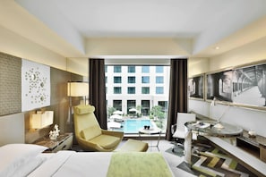 A hotel room with a bed, a yellow armchair, a desk with a lamp, a view of a swimming pool and building.