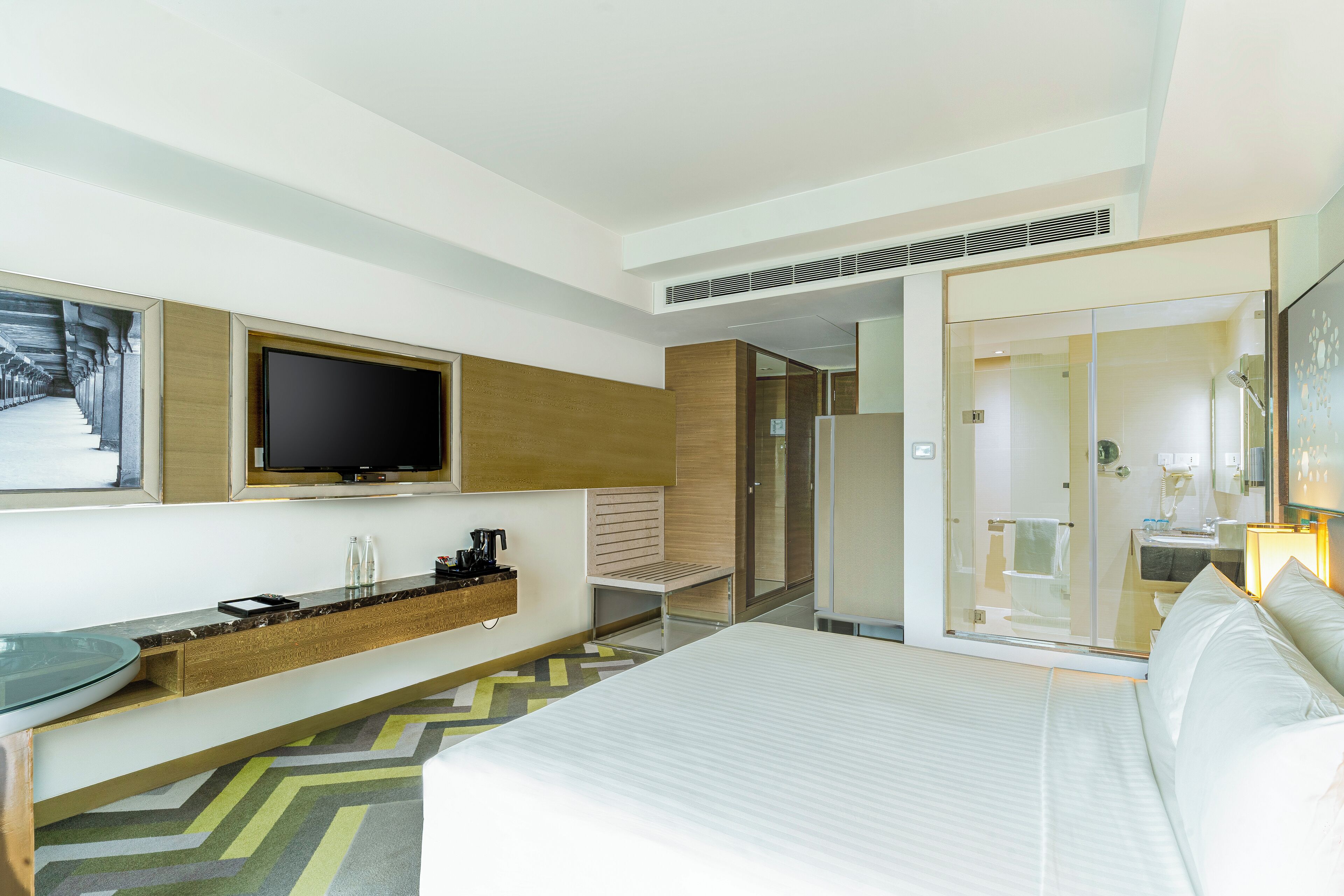 A modern hotel room with a large bed, a flat-screen TV mounted on the wall, a small seating area with a bench, and a shower area visible through a glass door.