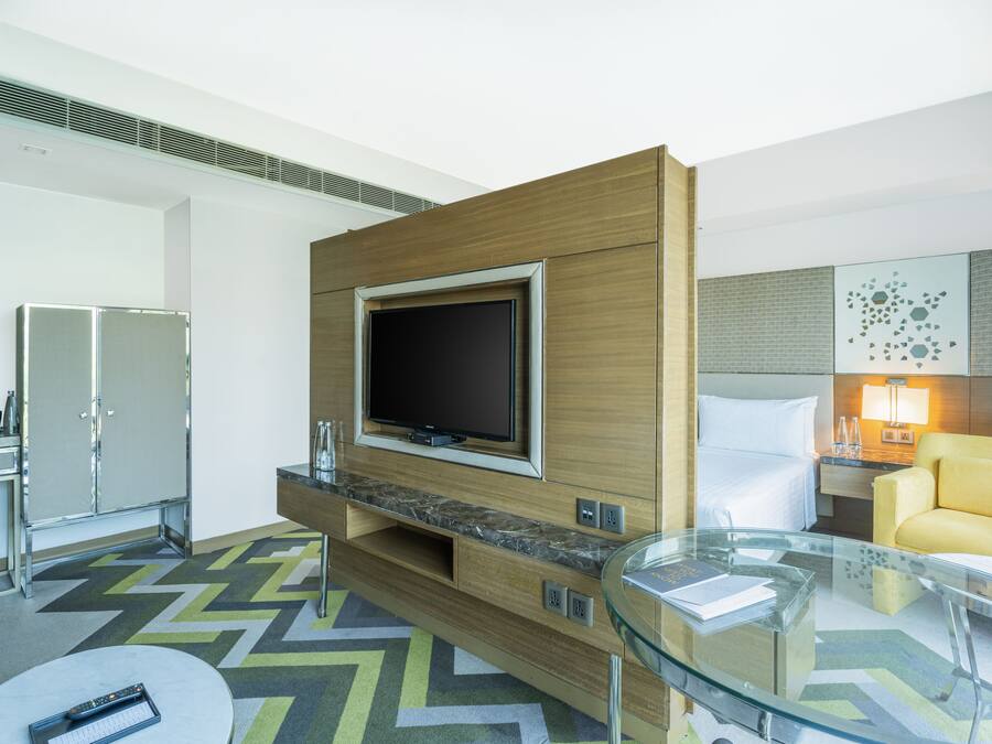 A modern hotel room with a large flat-screen TV mounted on a wooden panel, a glass coffee table, a yellow sofa, and a minibar.