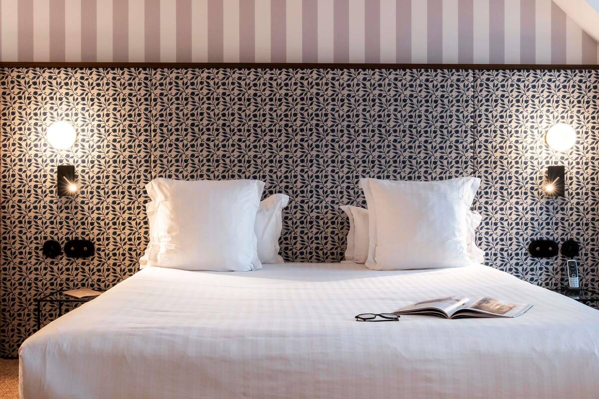A neatly made bed with white bedding, two bedside lamps, and a patterned wallpaper.