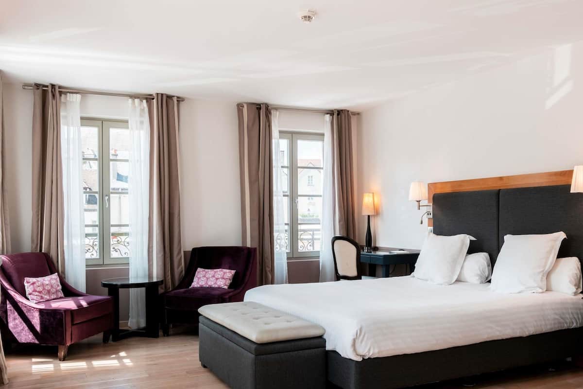 A hotel room with a large bed, two armchairs, a small table, and a window with curtains.