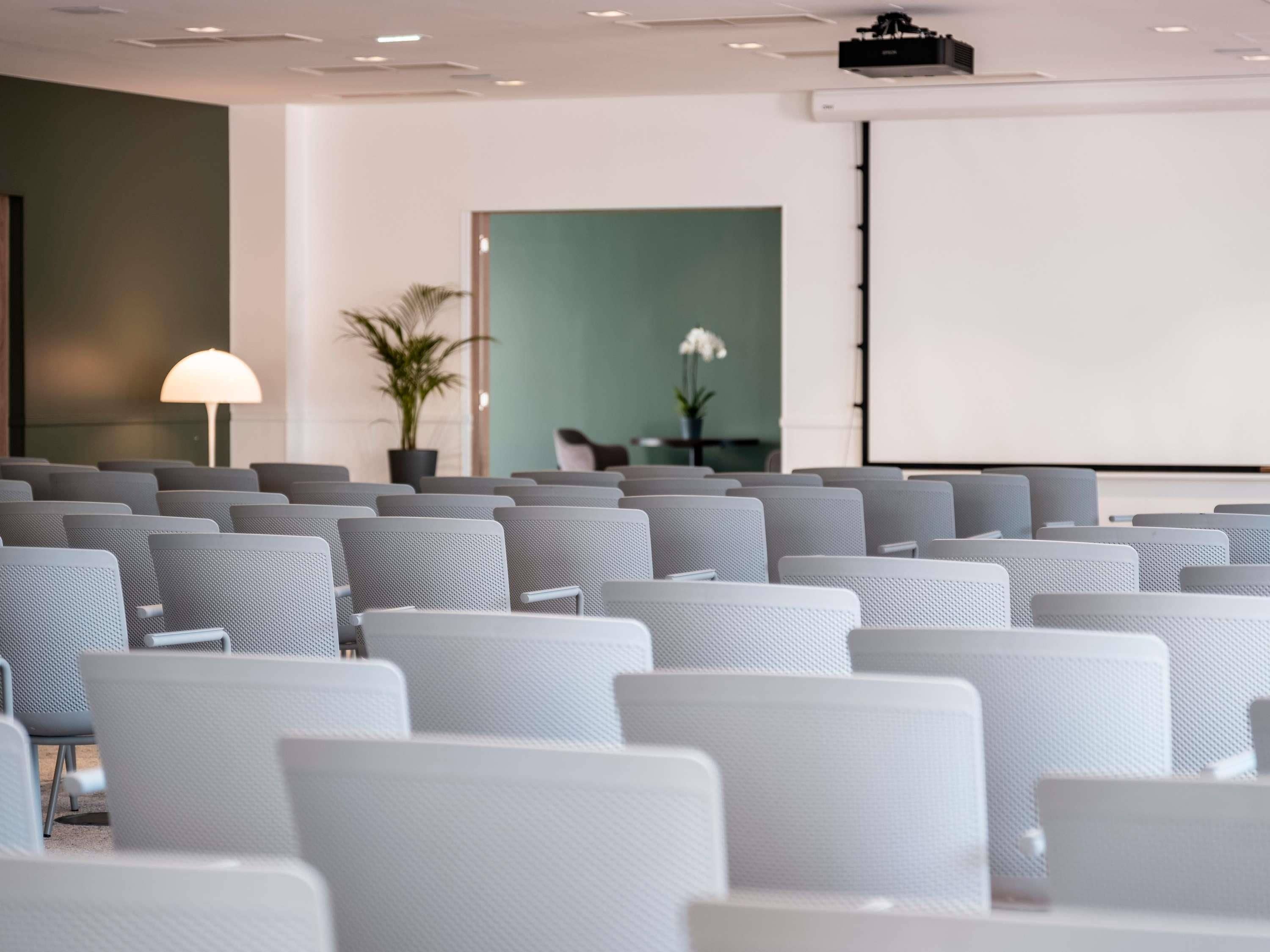 Ett konferensrum med rader av grå stolar, en projektor, en whiteboard och en krukväxt.