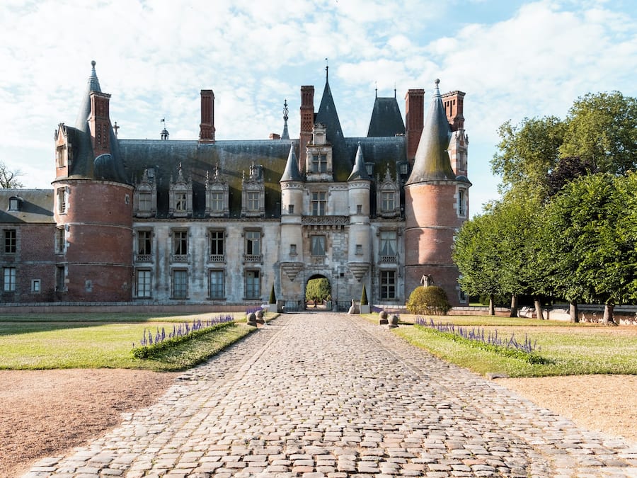 Um edifício histórico com um caminho de pedras que leva a uma entrada imponente, rodeado por jardins bem cuidados e árvores.