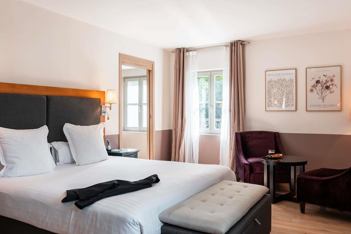 A hotel room with a large bed, a bedside table, a chair, a small table, and two framed pictures on the wall.