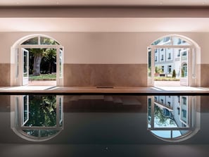 Un vestíbulo de hotel con piso reflectante, dos amplias ventanas en arco y vistas a un edificio y follaje exterior.