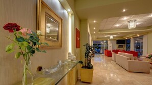 A hotel lobby with a reception desk, a large mirror, a vase with roses, and a seating area with red sofas. - Hotel Taxiarhis (Pineios)