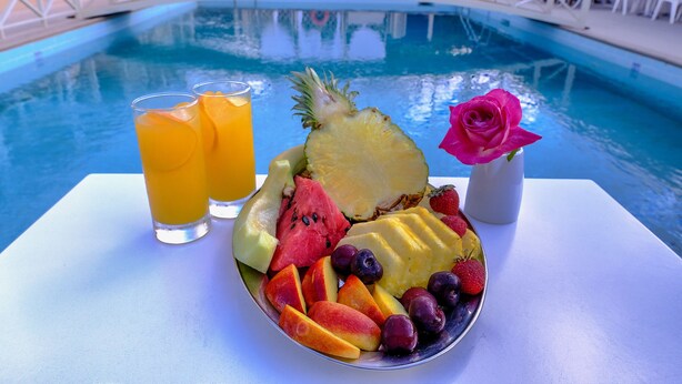 A poolside table with a fruit bowl, two glasses of orange juice, and a pink rose. - Hotel Taxiarhis (Pineios)
