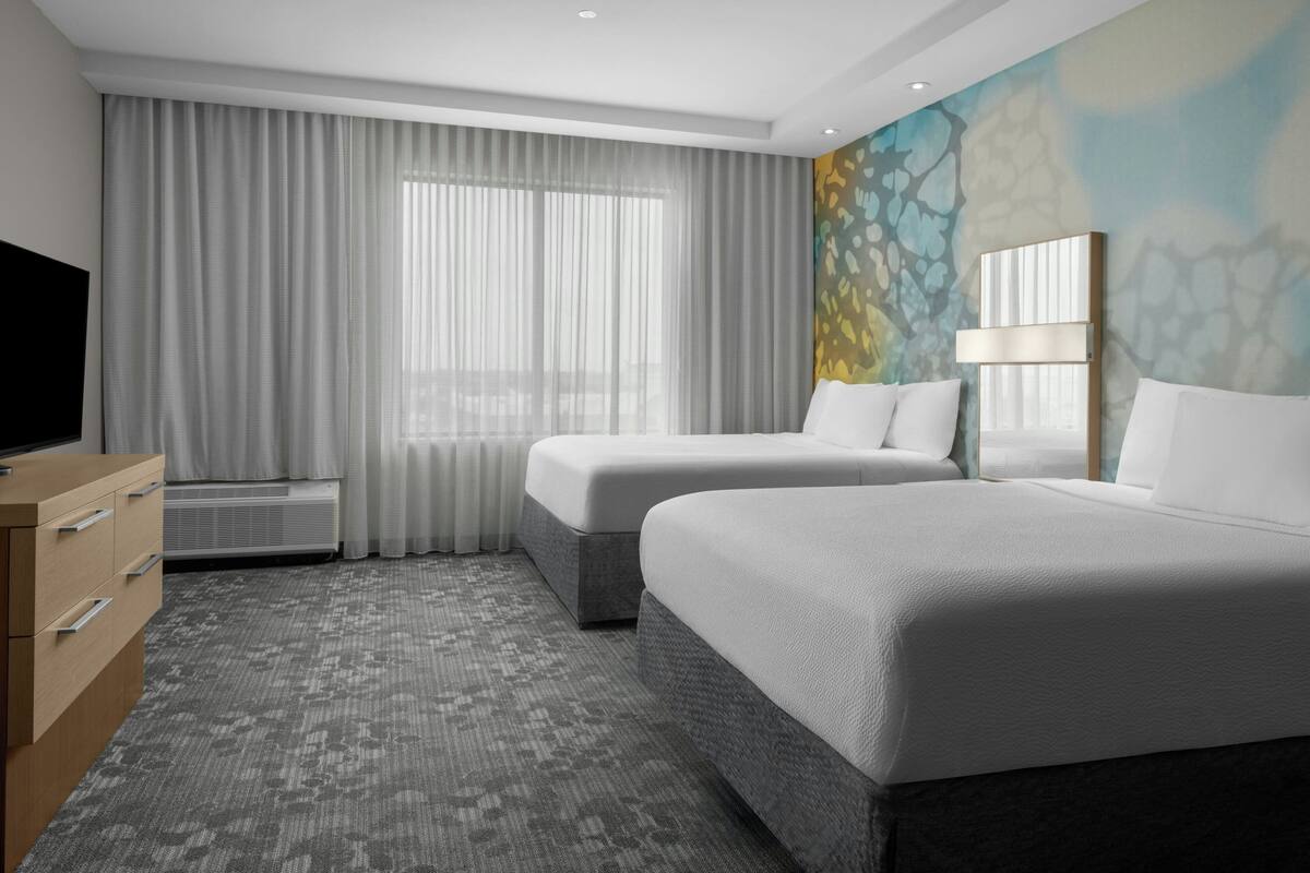 A hotel room with two beds, a TV, a dresser, and a large window with curtains.
