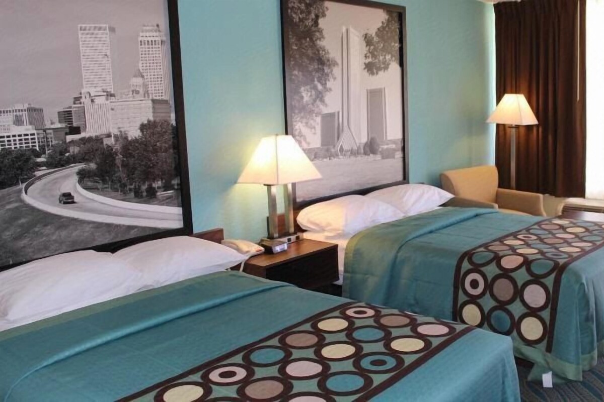 A hotel room with two beds, a nightstand with a lamp, a chair, and a window with curtains.