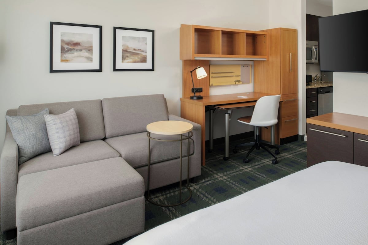 Habitación de hotel compacta con sofá, una pequeña mesa redonda, escritorio con silla, cama, kitchenette y dos cuadros en la pared.