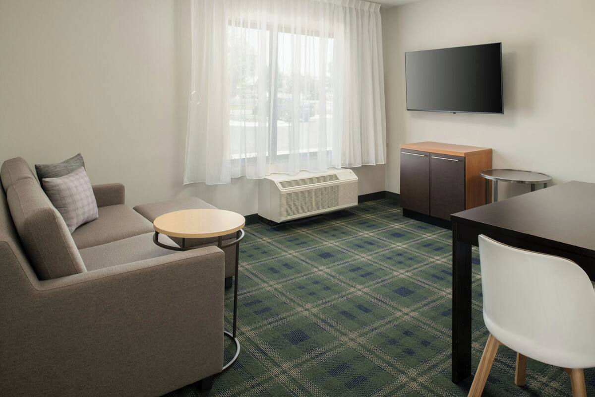 Habitación de hotel con sofá, una mesita redonda, una silla blanca, televisor montado en la pared y una pequeña zona de kitchenette.