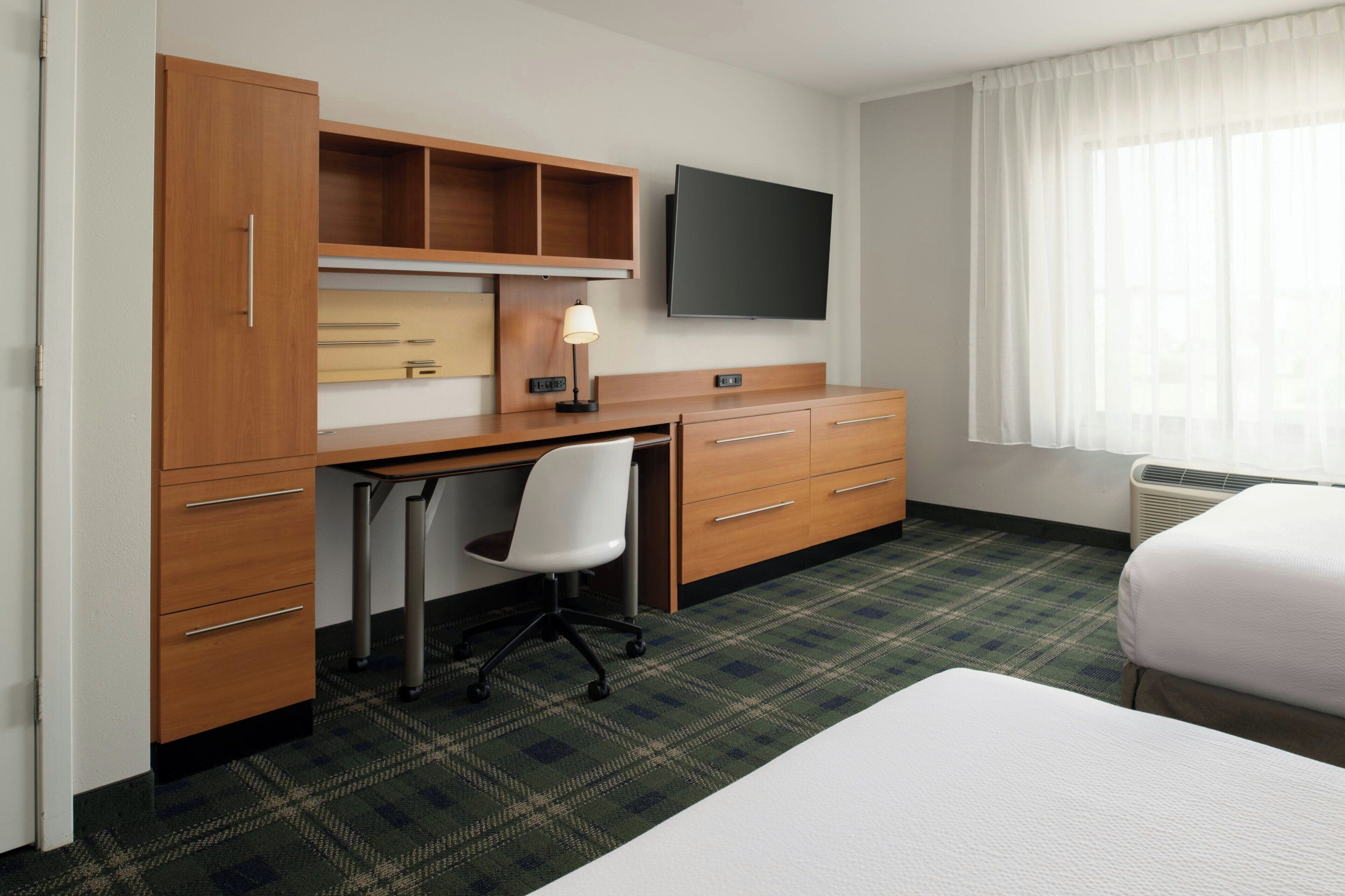 Habitación de hotel con escritorio de madera, televisor de pantalla plana, una silla blanca, cama y ventana con cortinas translúcidas.
