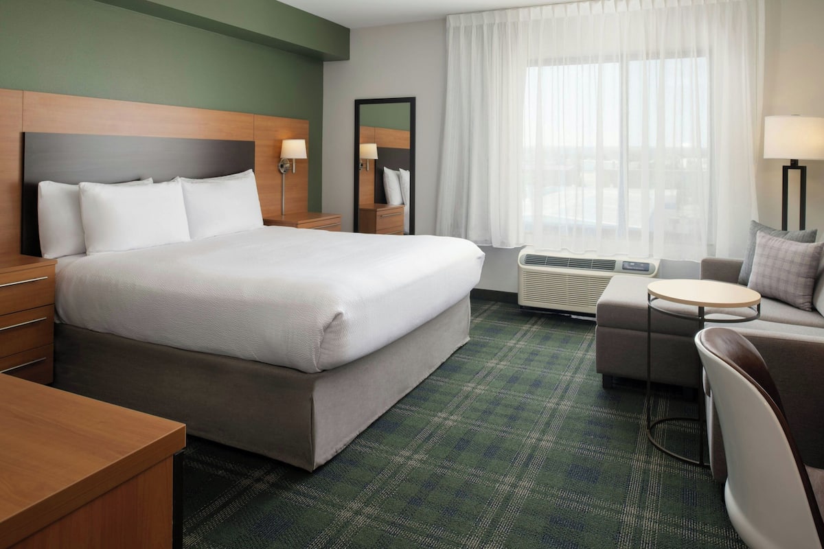 Habitación de hotel con una cama grande, una mesita pequeña, un sofá, un espejo y una ventana con cortinas.