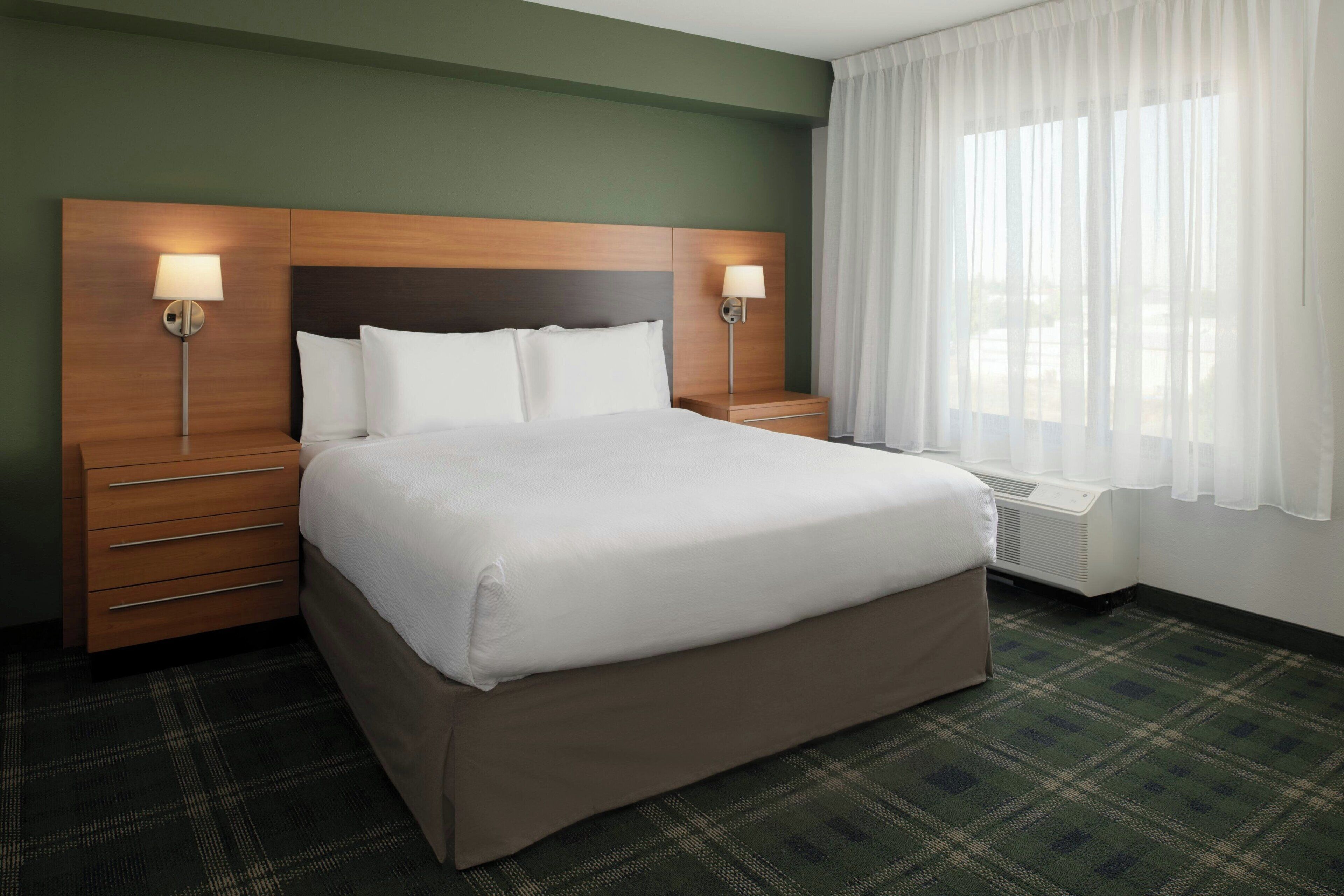 A hotel room with a large bed, two bedside tables with lamps, a green wall, and a window with sheer curtains.