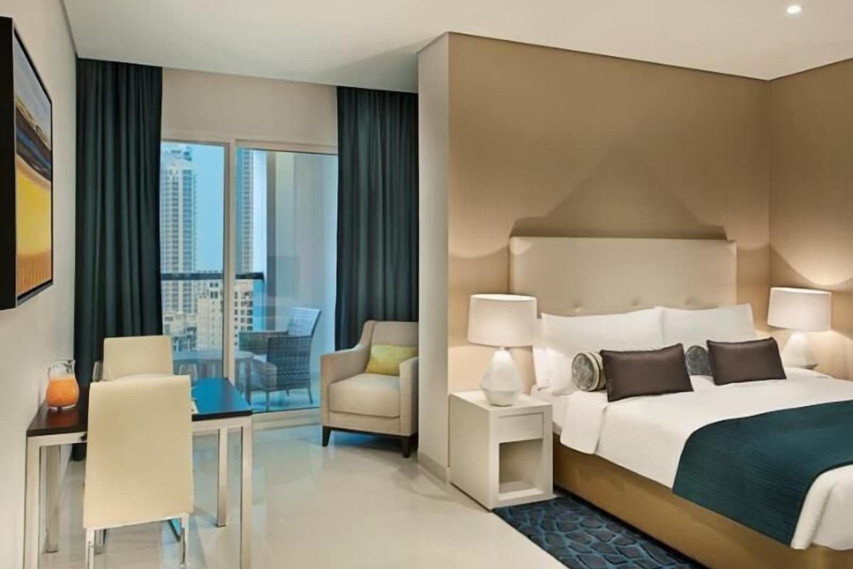 A modern hotel room with a large bed, bedside lamps, a desk with a chair, and a view of the city through large windows.