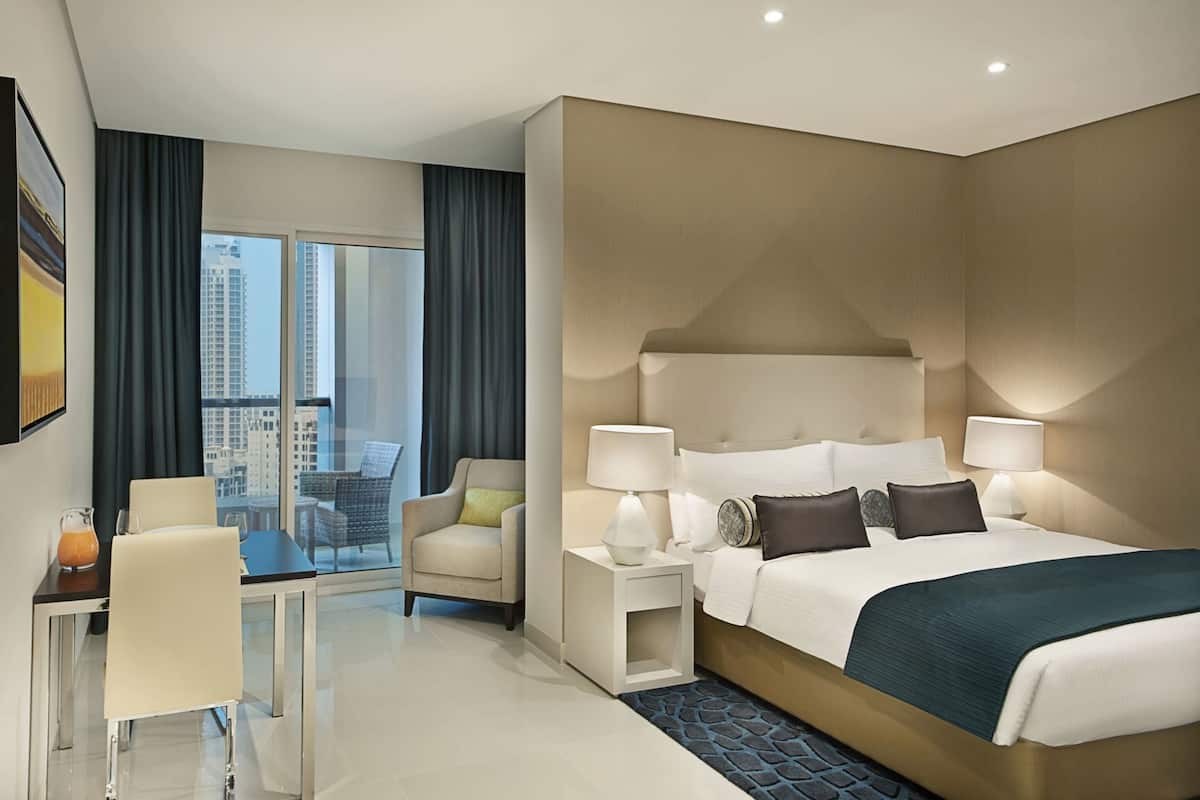 A hotel room with a large bed, bedside lamps, a chair, a small table, and a view of the city.