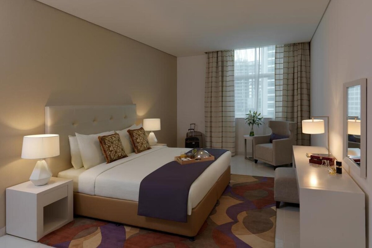 A hotel room with a bed, bedside tables, a desk, a chair, and a window with curtains.