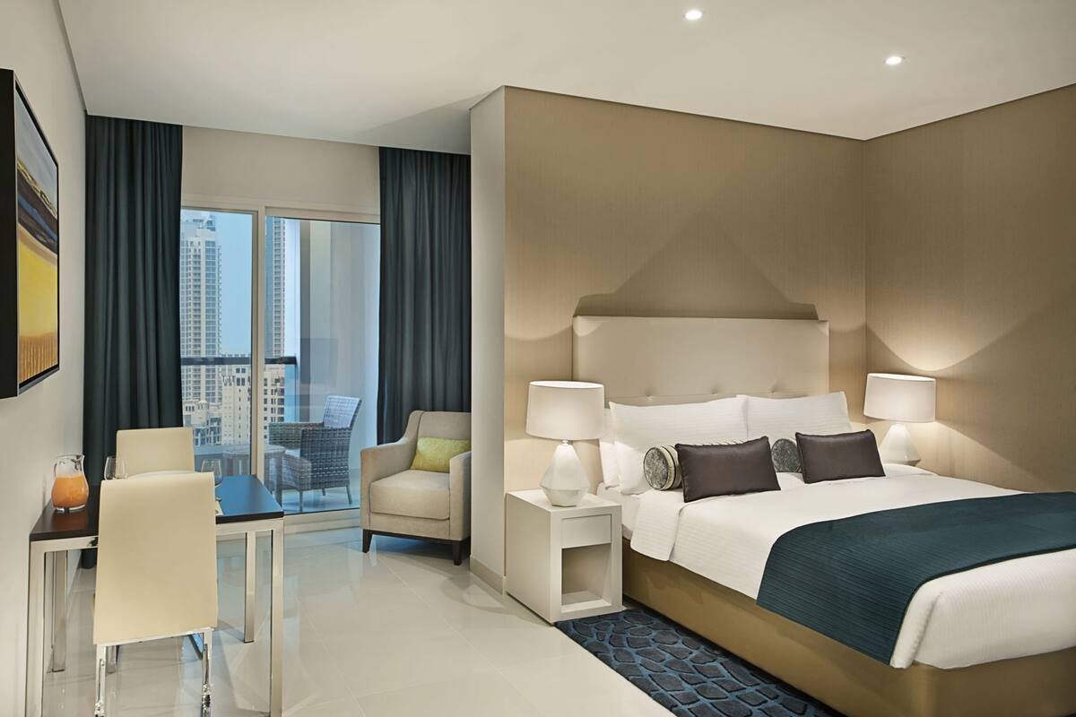 A hotel room with a large bed, bedside lamps, a chair, a small table, and a view of the city.