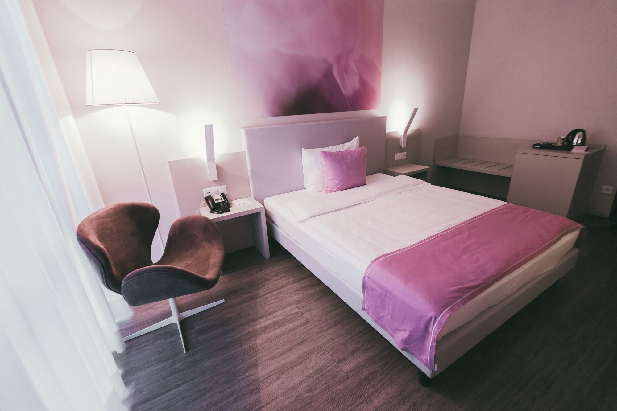 A modern hotel room with a bed, a desk, a chair, a phone, and a wall-mounted lamp.