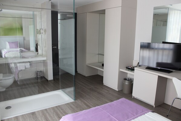 A modern hotel room with a glass shower enclosure, a white vanity, a bed with a purple blanket, and a flat-screen TV. - Hotel City Lugano, Design & Hospitality (Lugano)