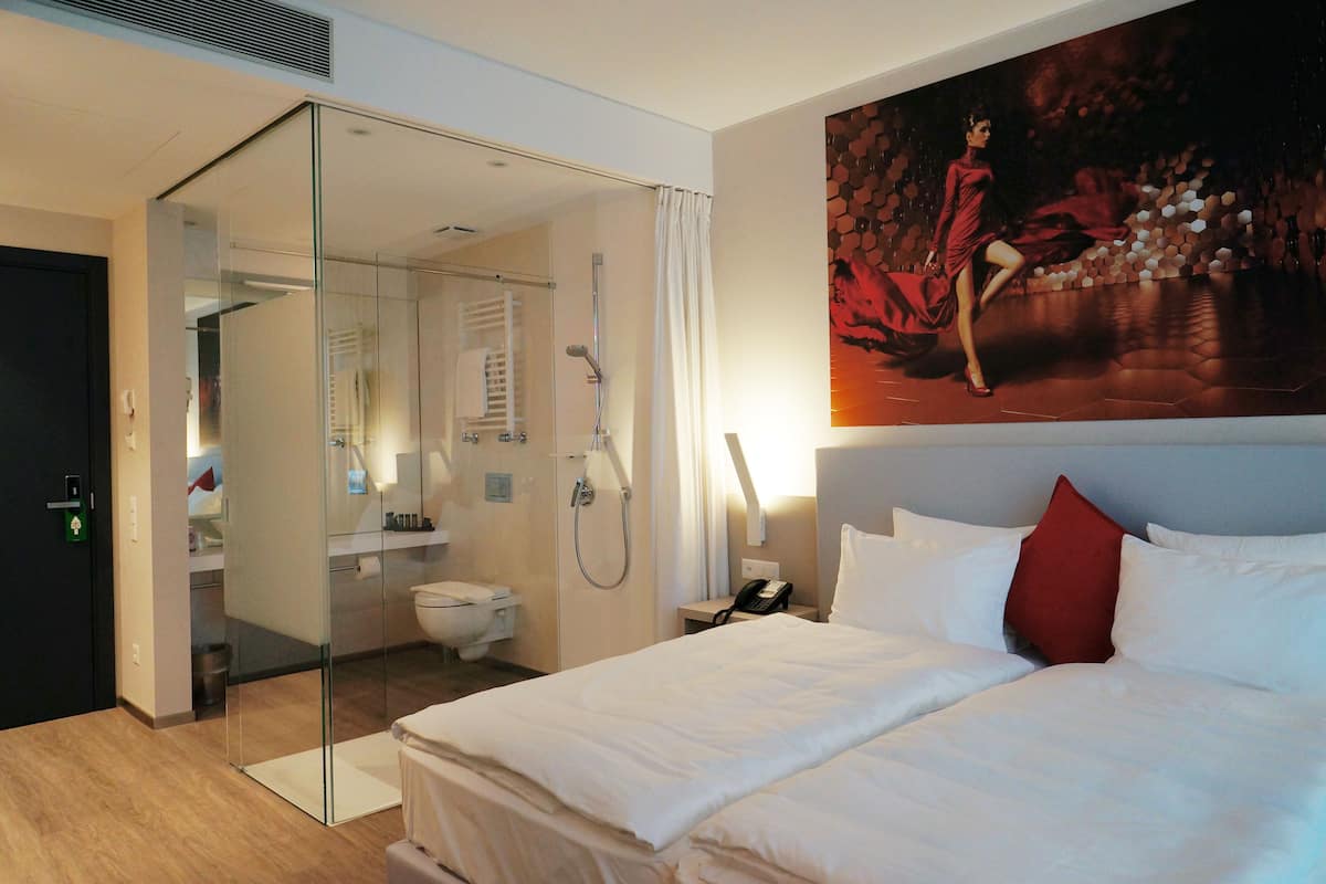 A hotel room with a bed, a glass shower enclosure, a bathroom with a toilet and sink, and a large painting on the wall.