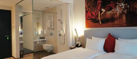 Habitación de hotel con cama, cabina de ducha de vidrio, baño con inodoro y lavamanos, y un cuadro grande en la pared.