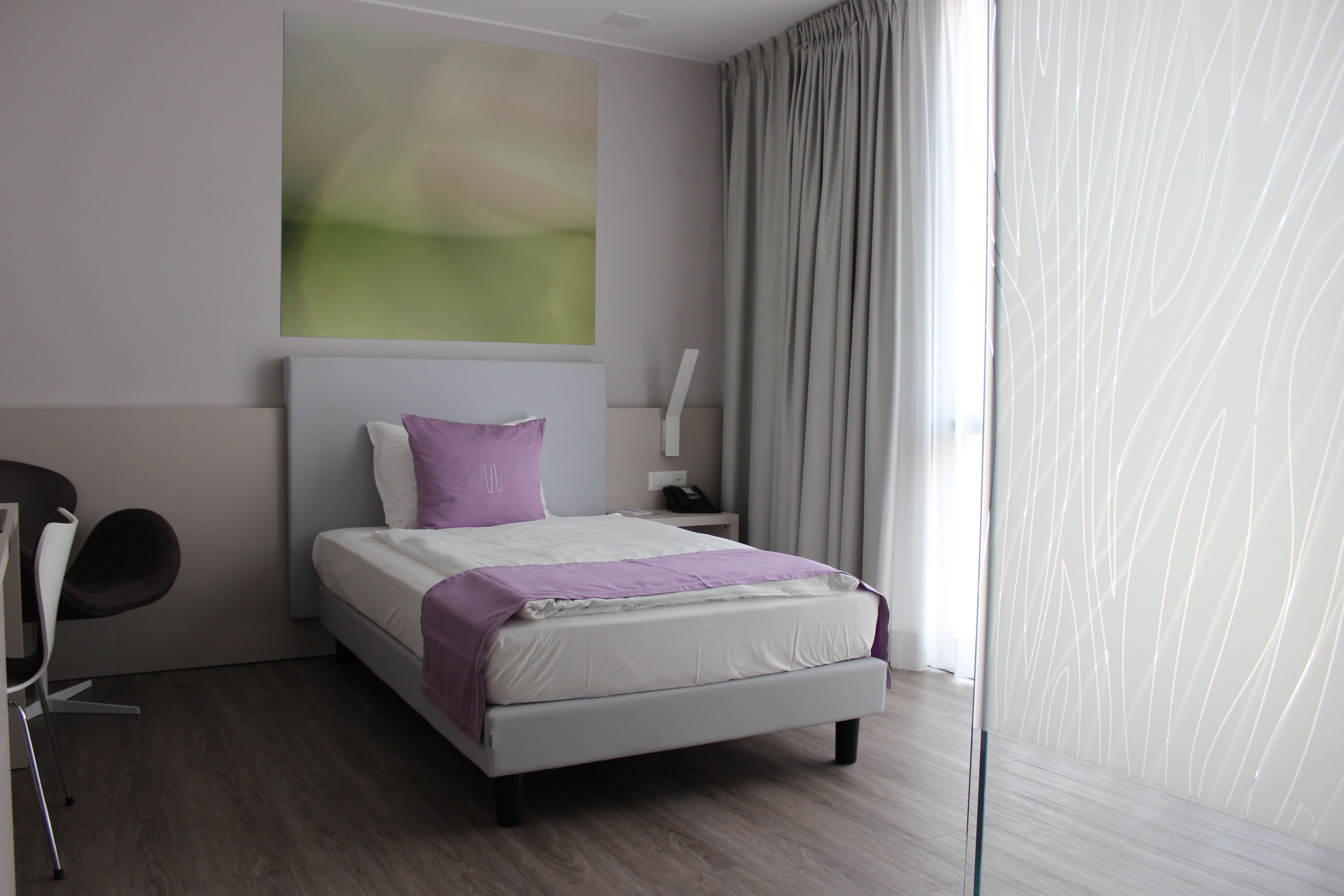 Ein modernes Hotelzimmer mit einem großen Bett, einem Nachttisch, einem Sessel und einem großen Fenster mit Vorhängen.