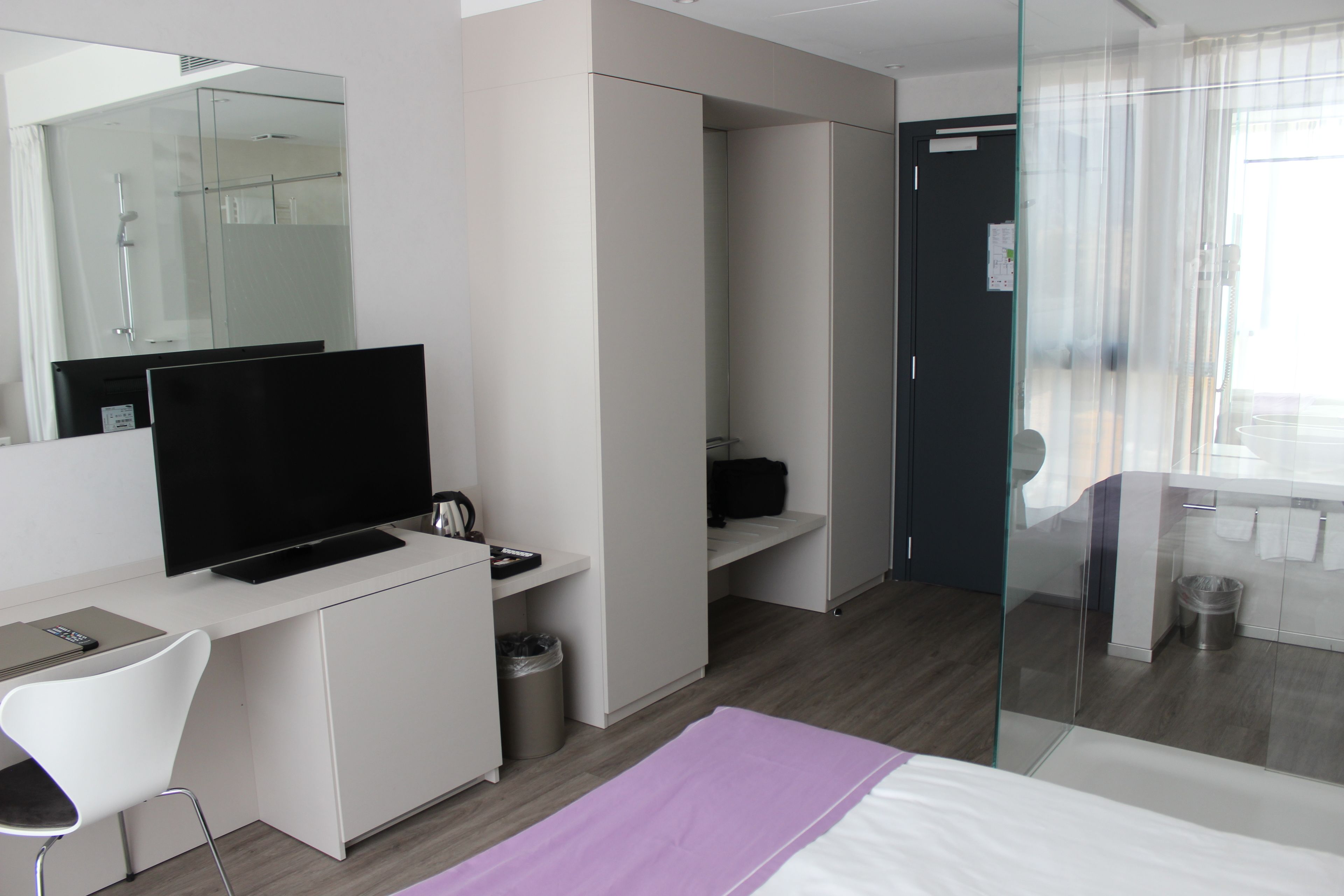 Una habitación de hotel moderna con una televisión de pantalla plana, un escritorio, una silla, una cama, cabina de ducha de vidrio y un pequeño cubo de basura.