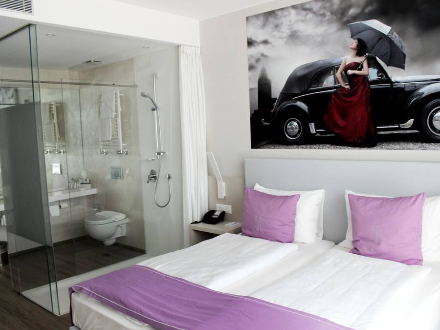 A modern hotel room with a glass shower enclosure, a bed with white and purple bedding, and a wall-mounted painting of a vintage car.
