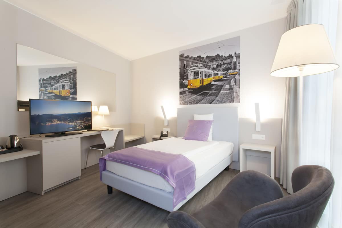 A modern hotel room with a bed, a desk, a chair, a television, and artwork on the walls.