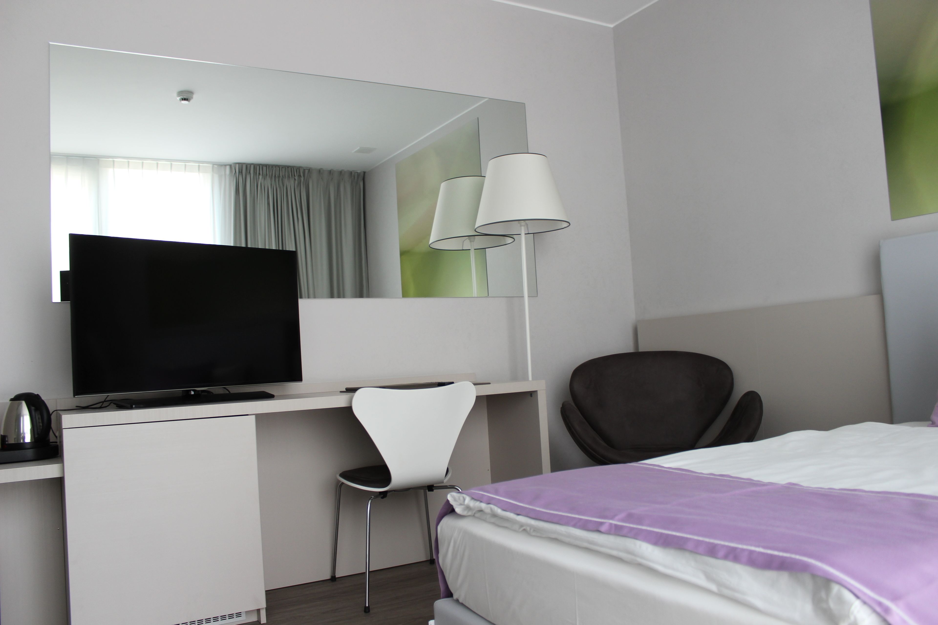 Una habitación de hotel moderna con una televisión de pantalla plana, un escritorio con una silla, un espejo grande y una cama con una colcha morada.