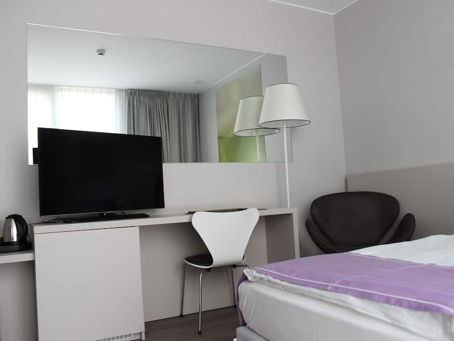 A modern hotel room with a flat-screen TV, a desk with a chair, a large mirror, and a bed with a purple blanket.