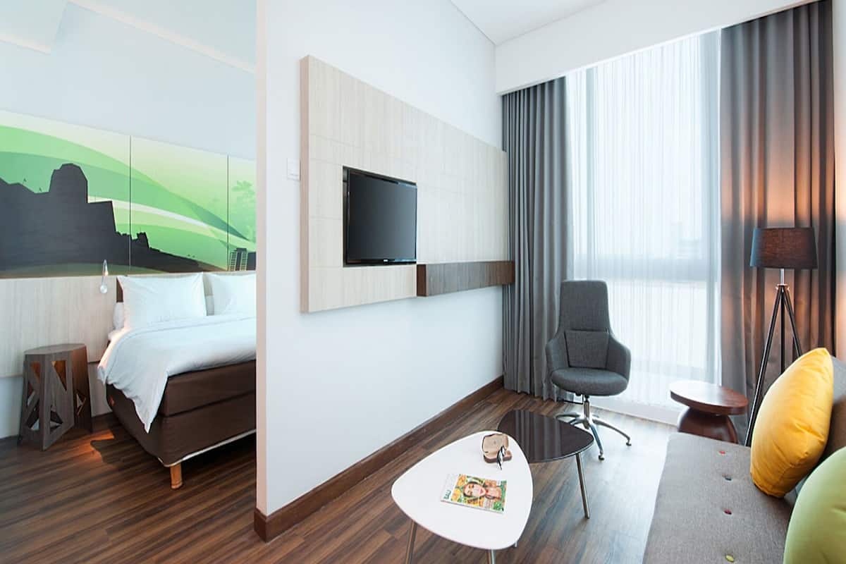 A hotel room with a bed, a TV mounted on the wall, a desk with a chair, a lamp, and a large window with curtains.