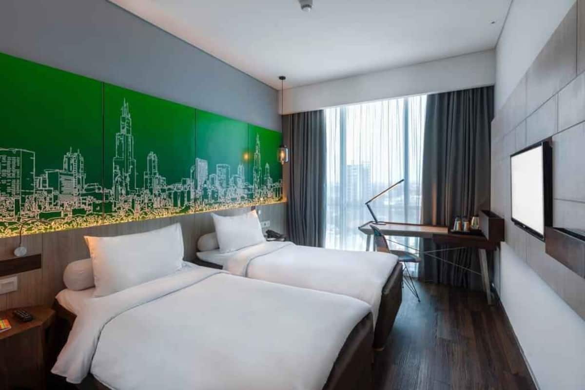A hotel room with two beds, a cityscape mural, a desk with a lamp, and a large window with curtains.